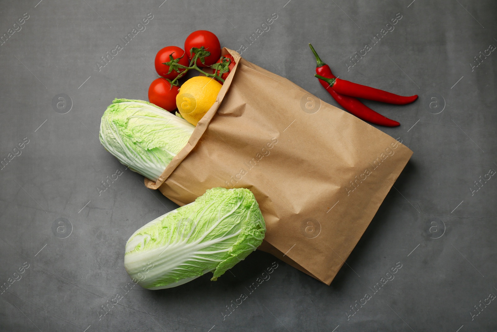 Photo of Paper bag with fresh Chinese cabbages, lemon, tomatoes and chili pepper on grey textured table, flat lay