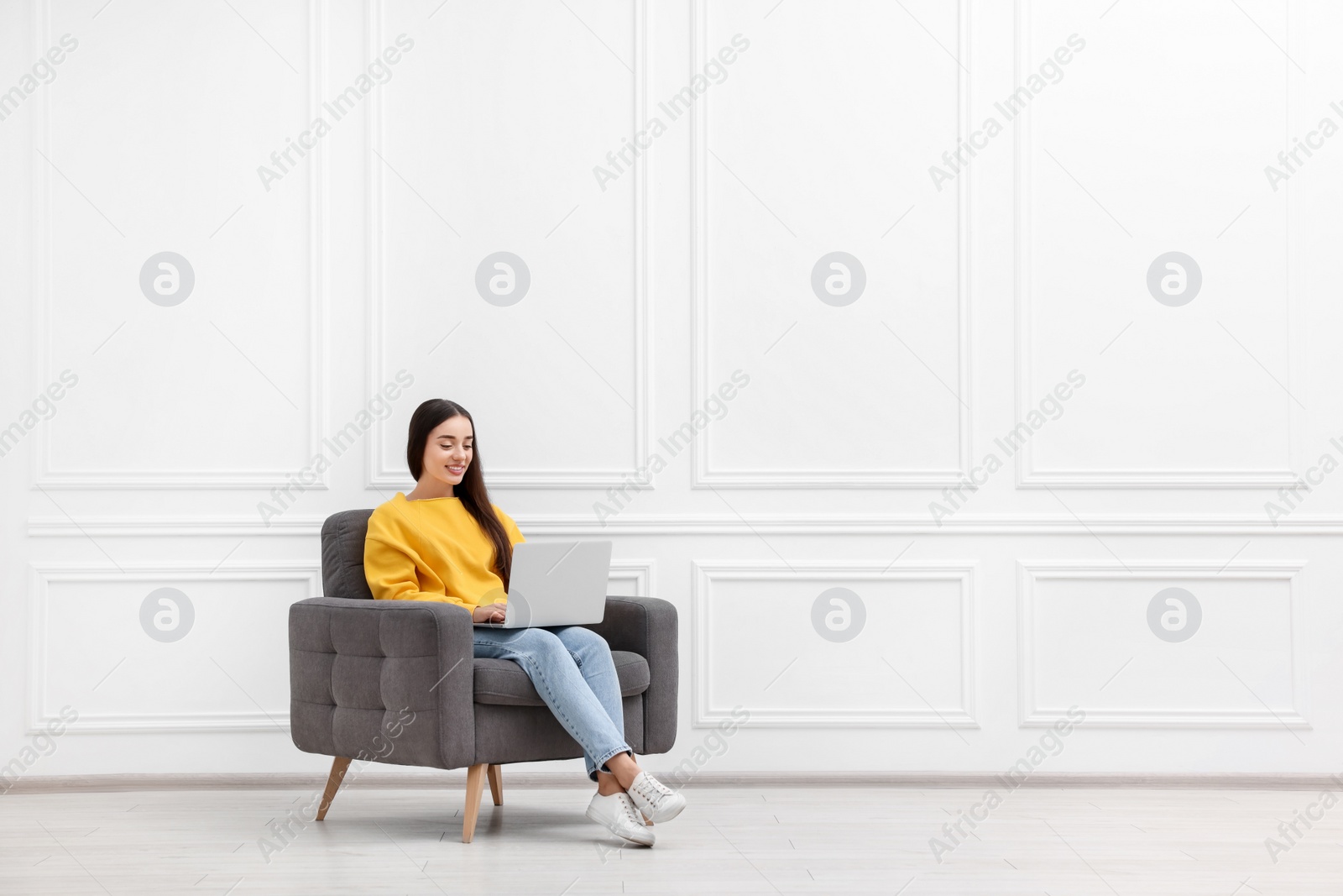 Photo of Beautiful woman with laptop sitting in armchair near white wall indoors, space for text