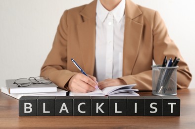 Black cubes with word Blacklist and woman working at wooden office desk, closeup