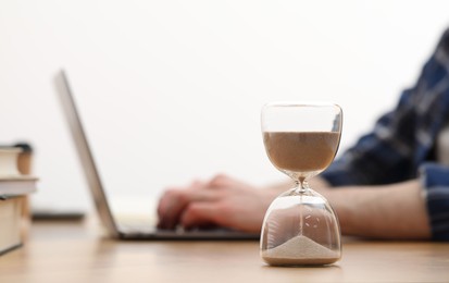 Photo of Hourglass with flowing sand on desk. Man using laptop indoors, selective focus