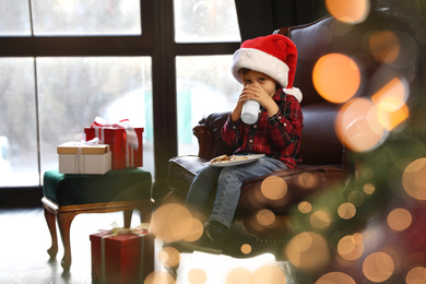 Photo of Little boy in Santa Claus cap with milk and cookies near window indoors. Christmas holiday