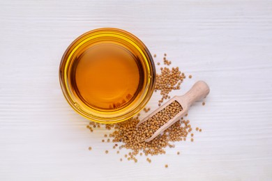 Photo of Bowl of natural oil and scoop with mustard seeds on white wooden table, flat lay