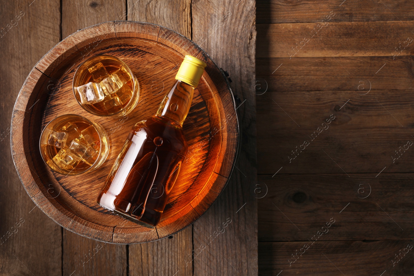 Photo of Whiskey with ice cubes in glasses, bottle and barrel on wooden table, top view. Space for text