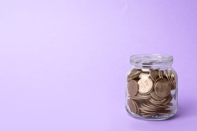 Glass jar with coins on lilac background, space for text