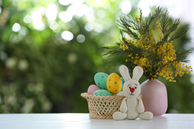 Photo of Easter bunny toy, dyed eggs and flowers on white wooden table against blurred green background. Space for text