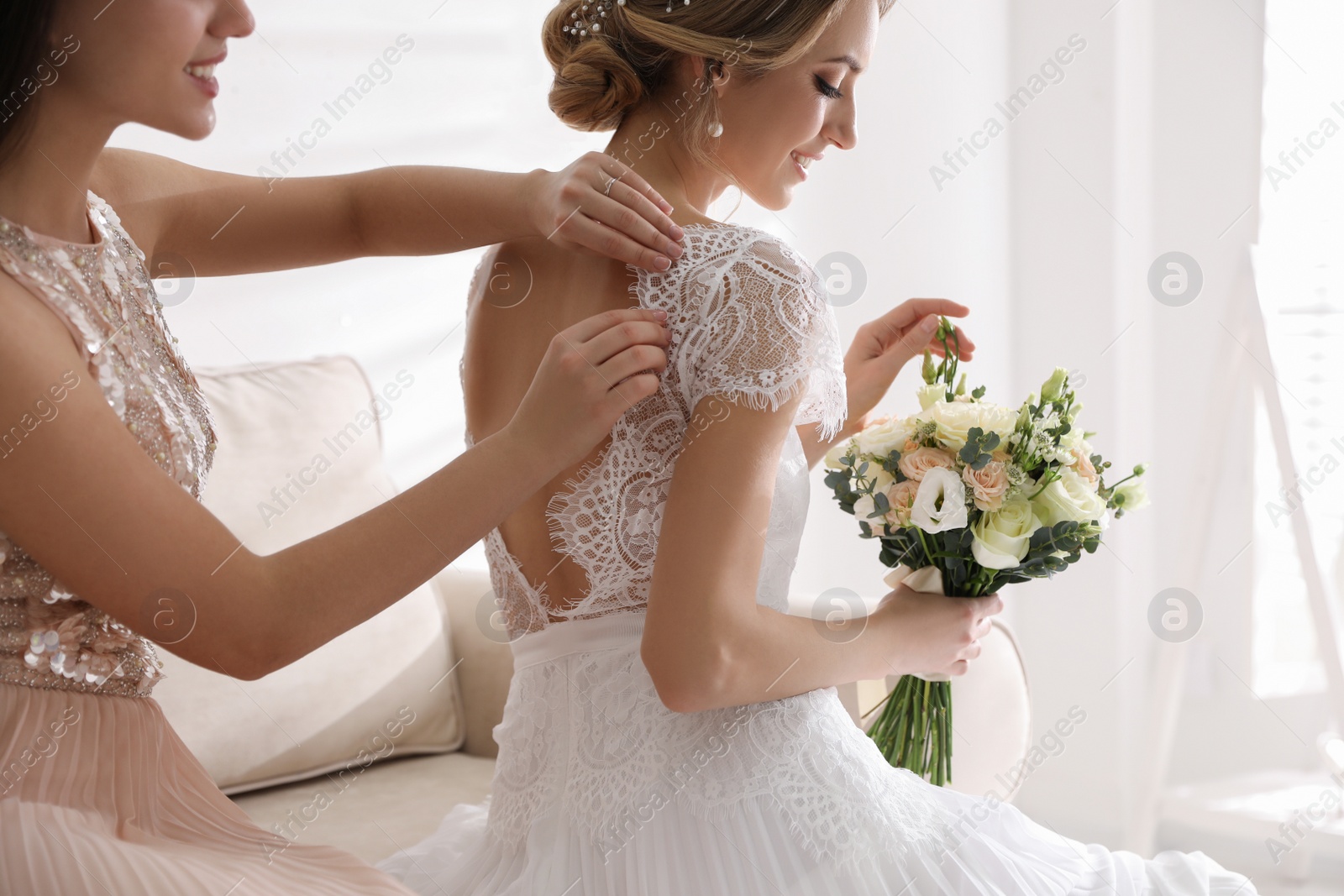 Photo of Young woman helping bride to put on wedding dress in room