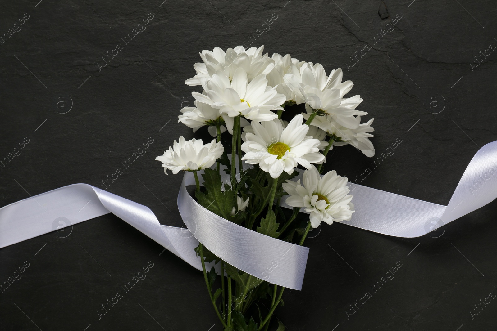 Photo of Beautiful chrysanthemum flowers and white ribbon on black table, top view. Funeral symbols