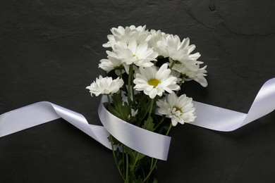 Beautiful chrysanthemum flowers and white ribbon on black table, top view. Funeral symbols