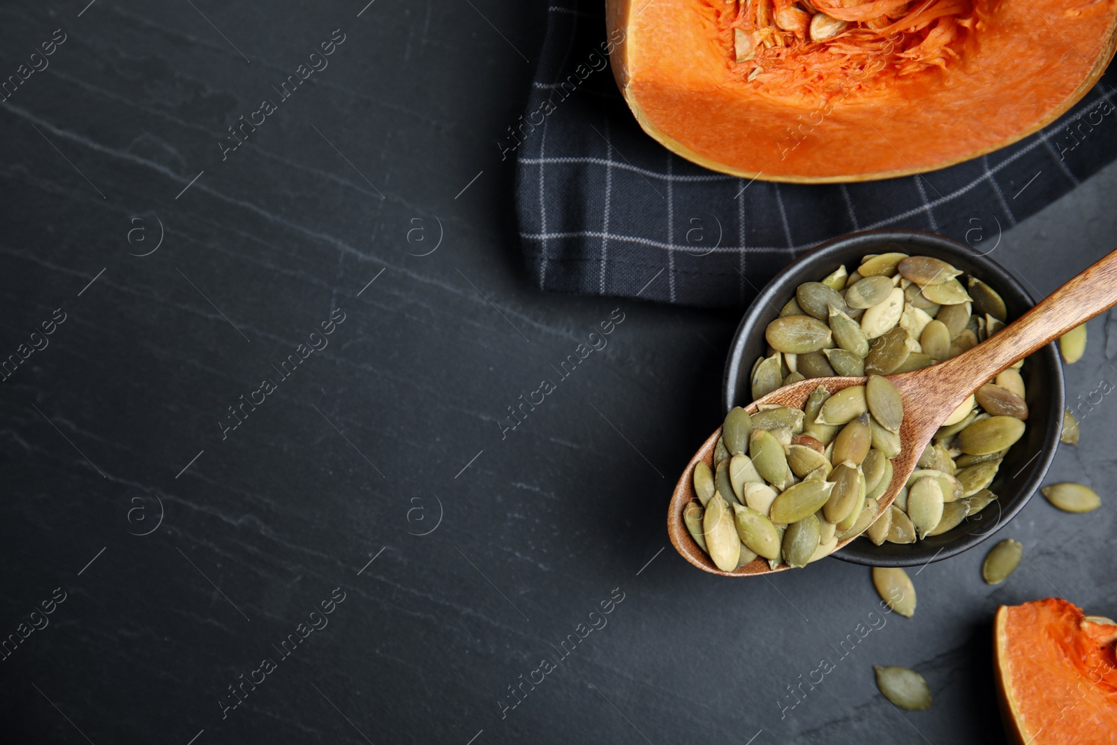 Photo of Bowl with seeds, wooden spoon and cut pumpkin on black table, flat lay. Space for text