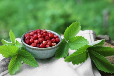 Bowl of tasty wild strawberries and green leaves on cloth against blurred background