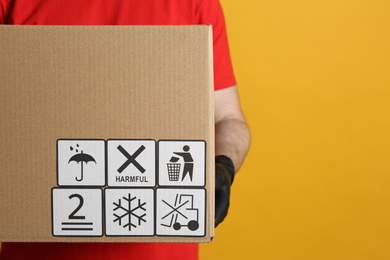 Photo of Courier holding cardboard box with different packaging symbols on yellow background, closeup. Parcel delivery