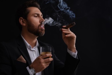 Photo of Handsome man in elegant suit with glass of whiskey smoking cigar on black background. Space for text
