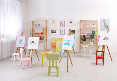 Photo of Easels with paintings and chairs for children in room