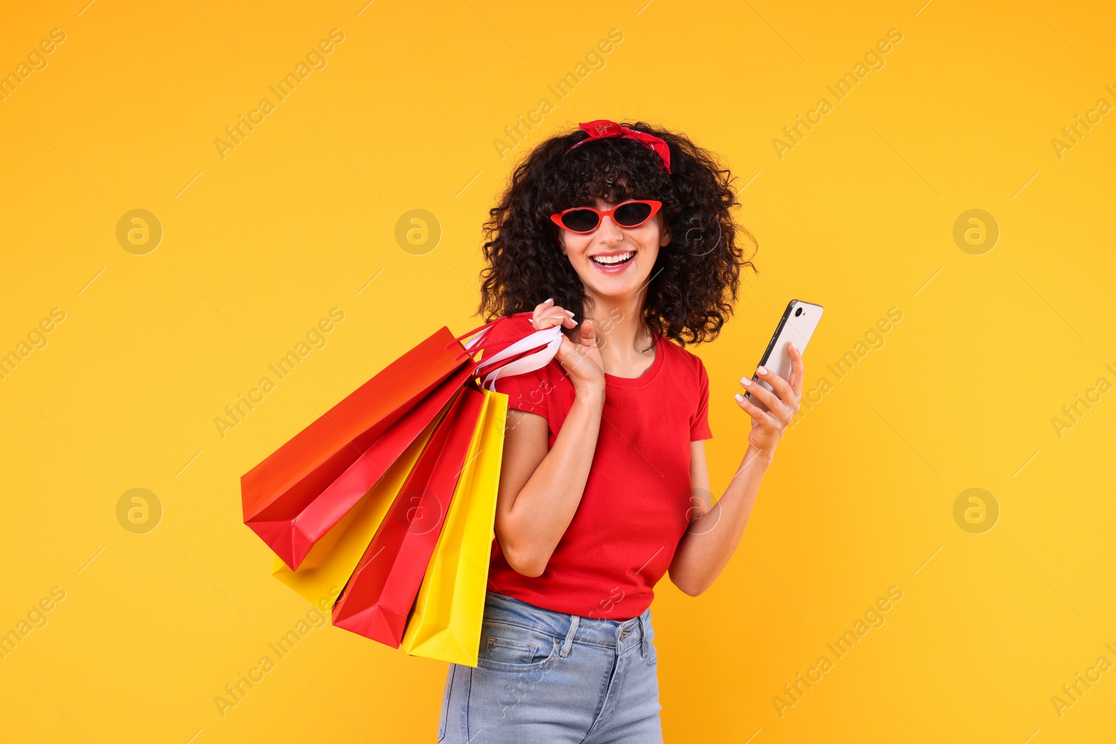 Photo of Happy young woman with shopping bags and modern smartphone on yellow background