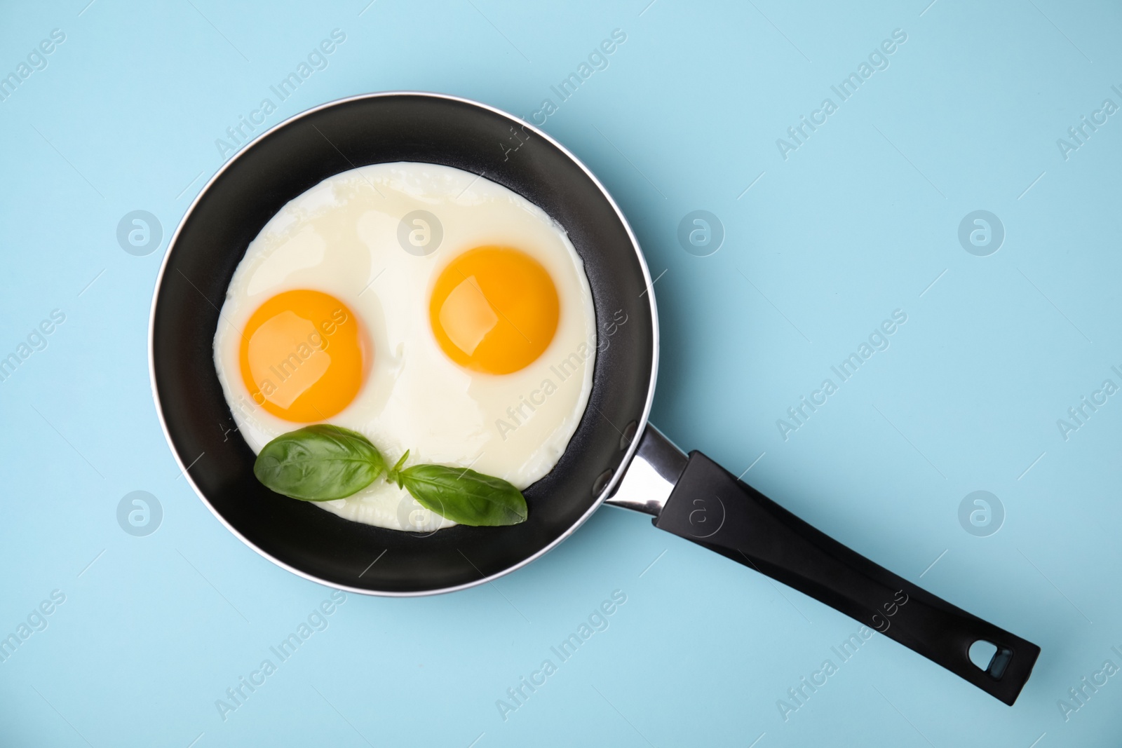 Photo of Tasty fried eggs with basil in pan on light blue background, top view