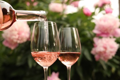Photo of Pouring rose wine from bottle into glass against beautiful peonies, closeup