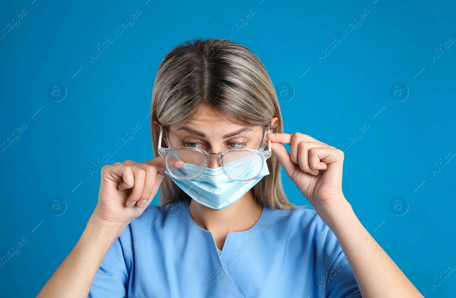 Photo of Doctor wiping foggy glasses caused by wearing disposable mask on blue background. Protective measure during coronavirus pandemic