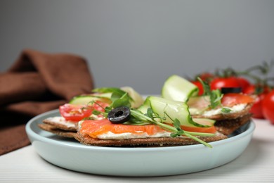 Photo of Tasty rye crispbreads with salmon, cream cheese and vegetables served on white wooden table, closeup