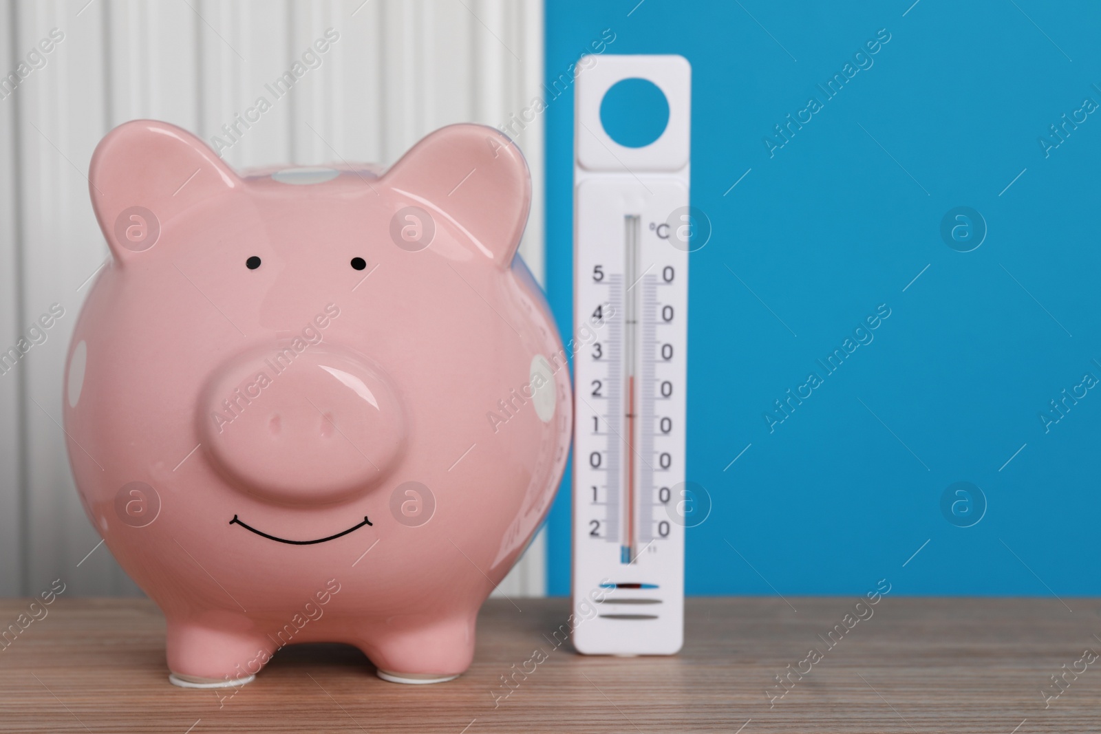 Photo of Piggy bank with thermometer on wooden table near heating radiator. Space for text