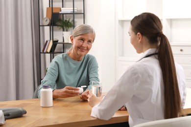 Photo of Young healthcare worker giving glass of water to senior woman with pills at wooden table indoors