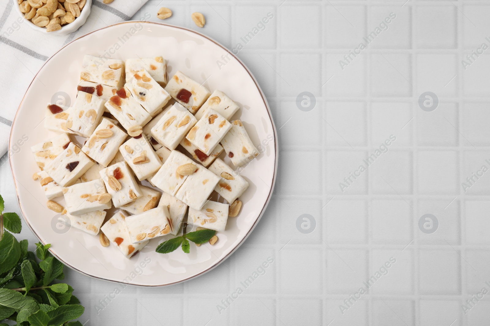 Photo of Pieces of delicious nutty nougat, peanuts and mint on white tiled table, flat lay. Space for text
