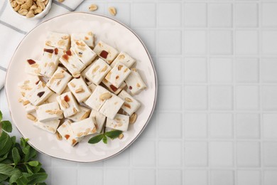 Pieces of delicious nutty nougat, peanuts and mint on white tiled table, flat lay. Space for text