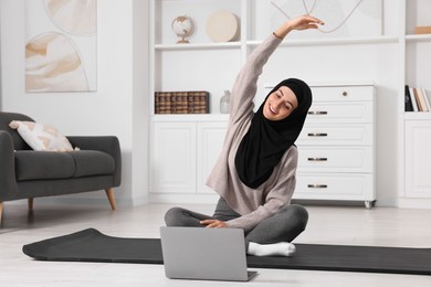 Photo of Muslim woman in hijab doing exercise near laptop on fitness mat at home