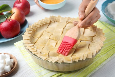 Woman spreading egg yolk onto raw apple pie at white wooden table, closeup