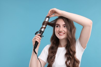 Photo of Beautiful young woman using curling hair iron on light blue background, space for text