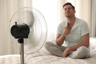 Photo of Man enjoying air flow from fan on bed in room, focus on device. Summer heat