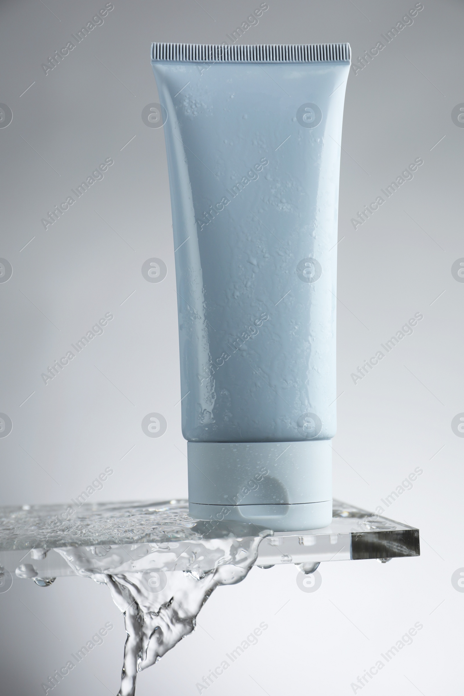 Photo of Moisturizing cream in tube on glass with water drops against grey background