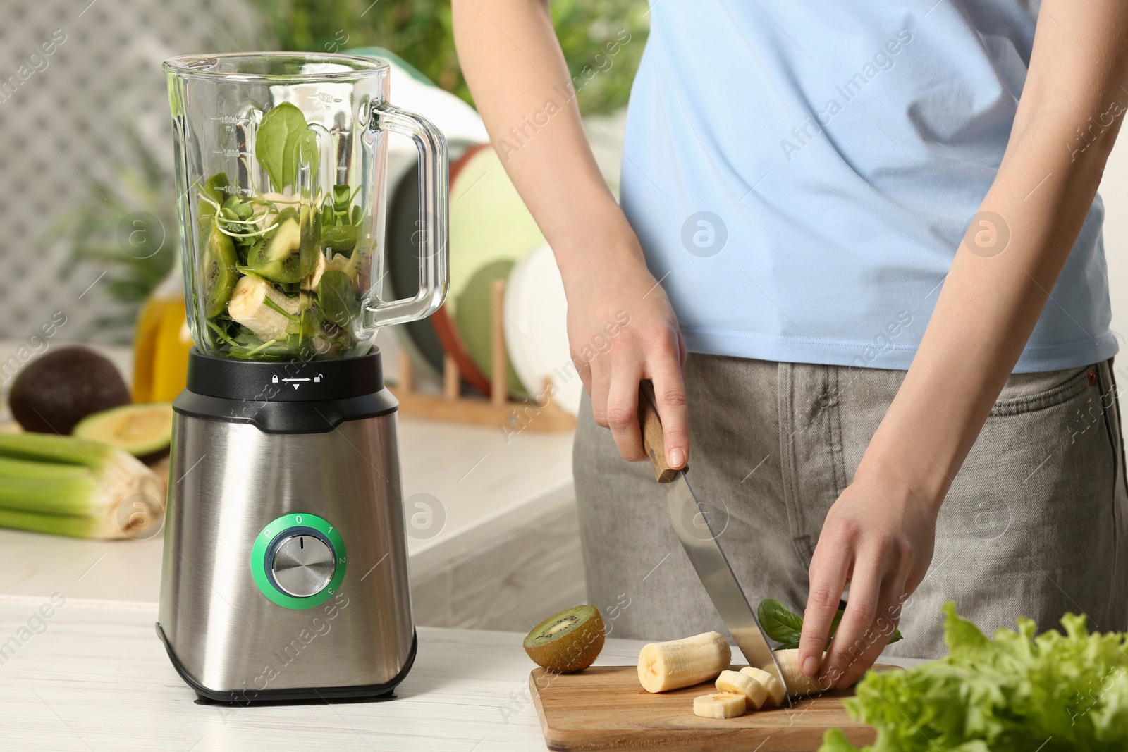 Photo of Woman preparing ingredients for tasty green smoothie at white wooden table in kitchen, closeup