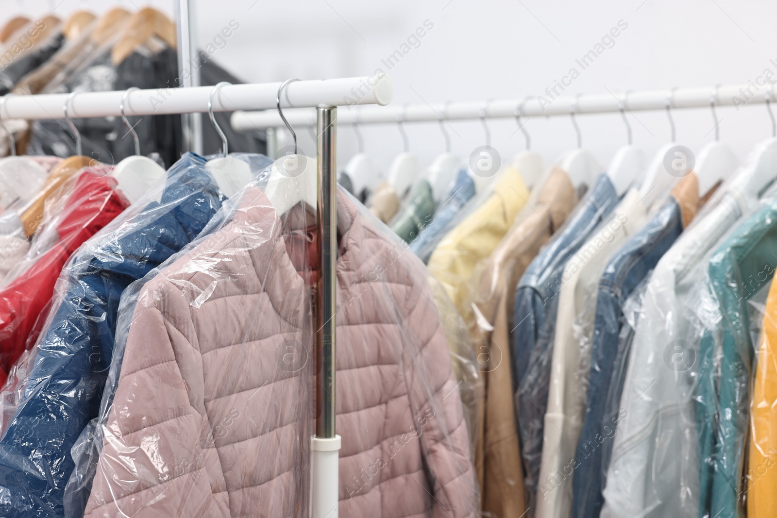 Photo of Dry-cleaning service. Many different clothes in plastic bags hanging on rack indoors, closeup