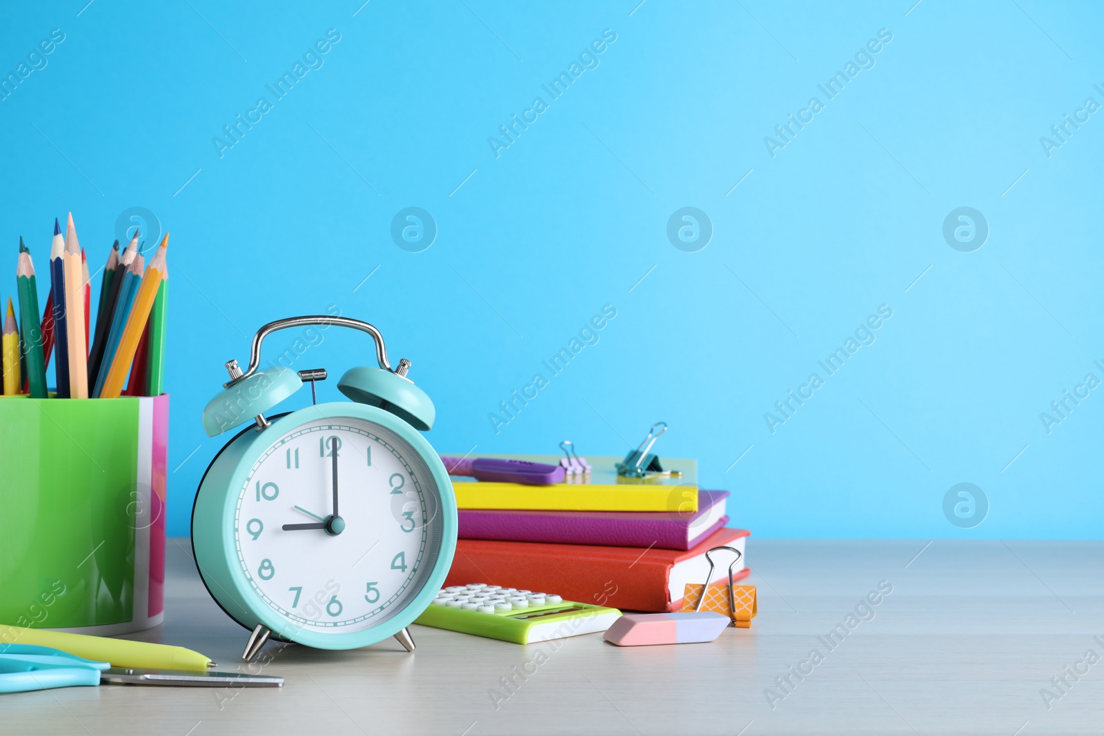 Photo of Turquoise alarm clock and different stationery on white wooden table against light blue background, space for text. School time