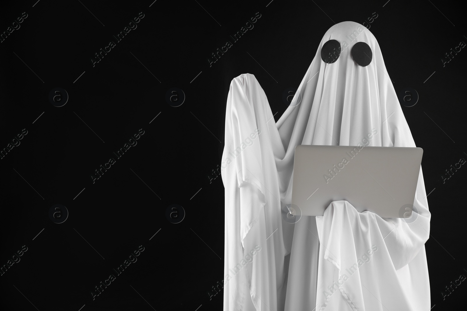 Photo of Creepy ghost. Person in white sheet with laptop on black background, space for text