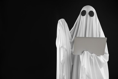 Photo of Creepy ghost. Person in white sheet with laptop on black background, space for text