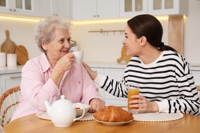 Photo of Young caregiver and senior woman having breakfast at table in kitchen. Home care service