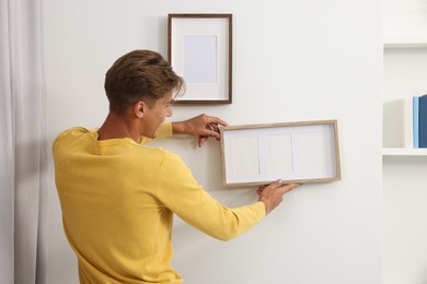 Photo of Young man hanging picture frames on white wall indoors, back view