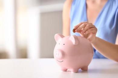 Photo of Woman putting coin into piggy bank at table indoors, closeup. Space for text