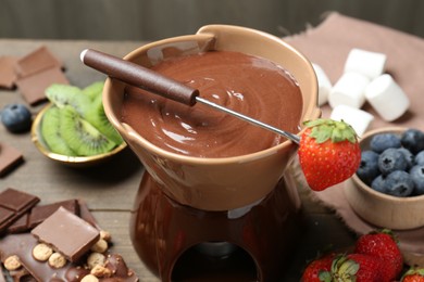 Fondue pot with melted chocolate, different fresh berries, kiwi and fork on wooden table, closeup