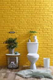 Photo of Stylish bathroom with toilet bowl, green plants and decor elements near yellow brick wall. Interior design
