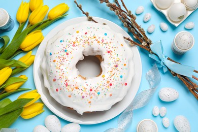 Delicious Easter cake decorated with sprinkles near beautiful tulips, willow branches and painted eggs on light blue background, flat lay
