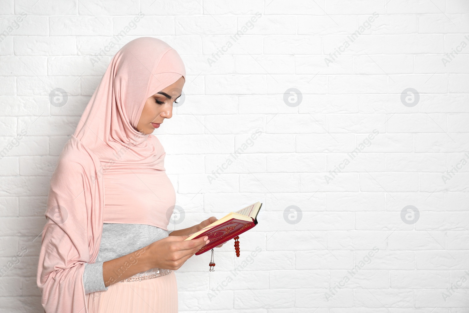 Photo of Young Muslim woman in hijab reading Koran against brick wall. Space for text