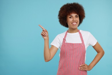 Photo of Happy young woman in apron pointing at something on light blue background. Space for text