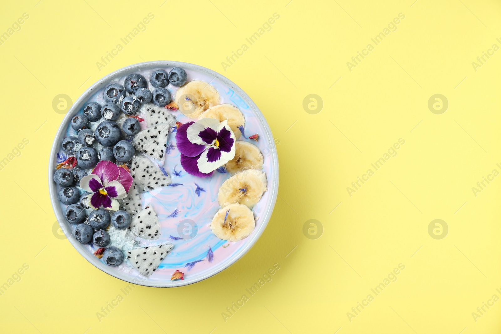 Photo of Delicious smoothie bowl with fresh fruits, blueberries and flowers on yellow background, top view. Space for text