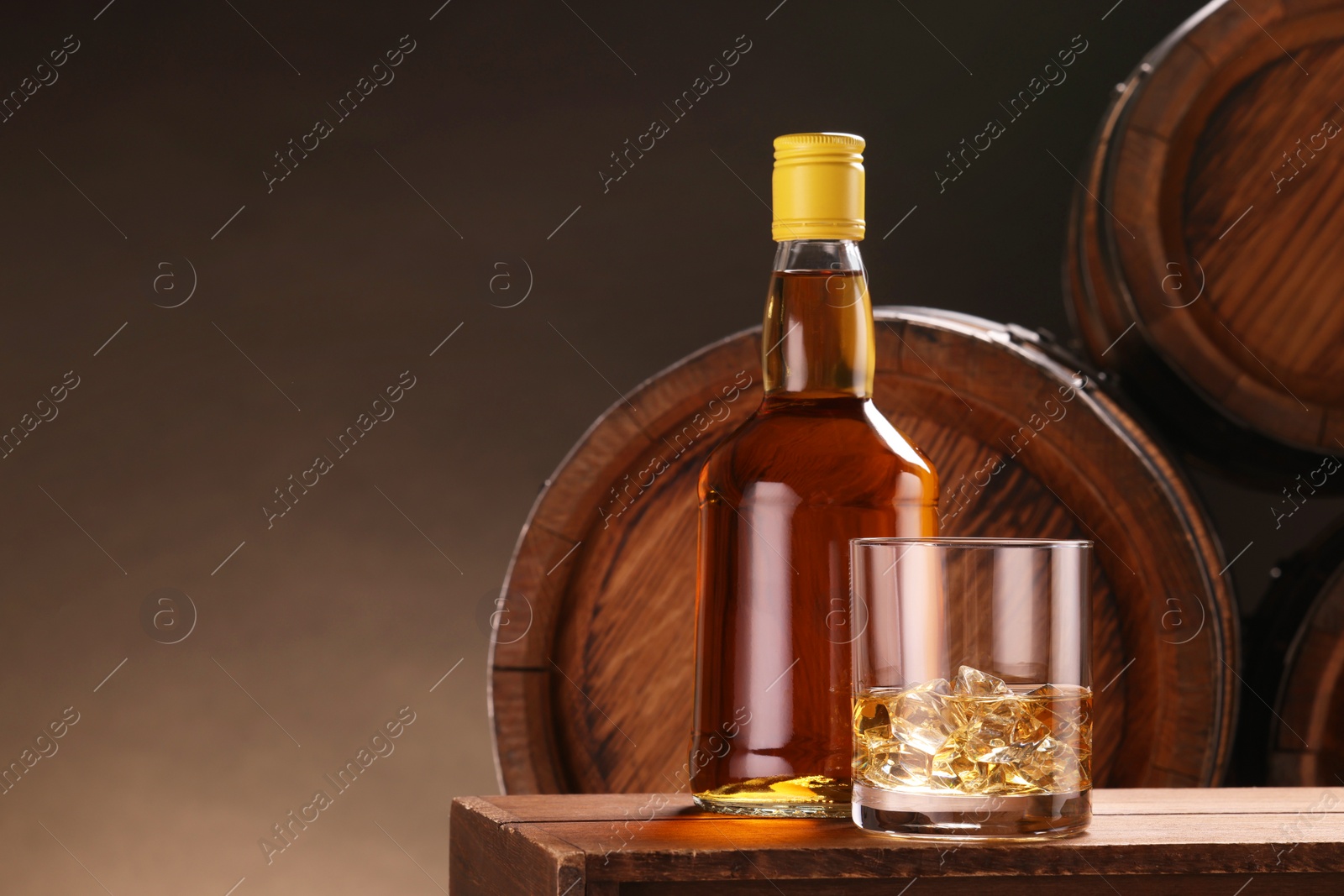 Photo of Whiskey with ice cubes in glass and bottle on wooden table near barrels against dark background, space for text