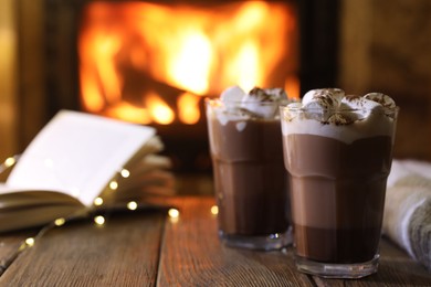 Photo of Glasses with hot cocoa, marshmallows, lights and book on wooden table near fireplace