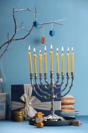 Hanukkah celebration. Menorah with burning candles, dreidels, donuts and gift boxes on table against light blue background, closeup