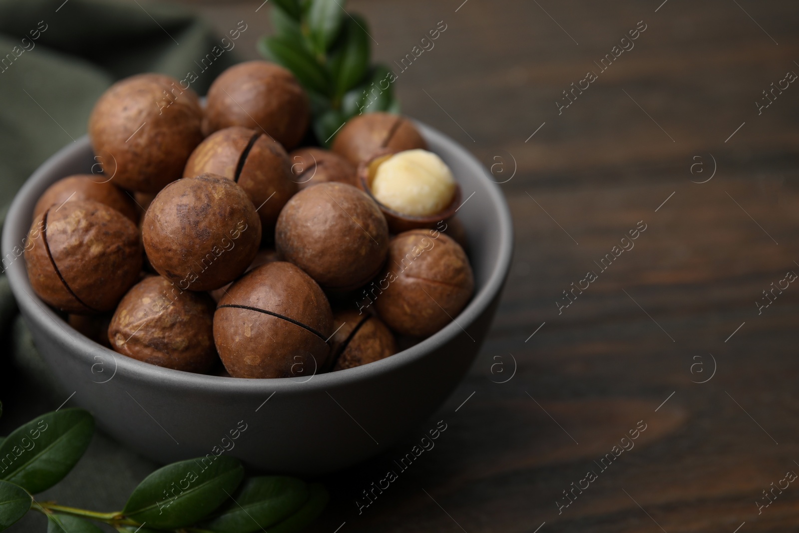 Photo of Tasty Macadamia nuts in bowl and green twigs on wooden table, closeup. Space for text