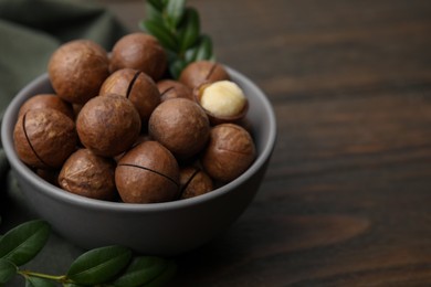 Photo of Tasty Macadamia nuts in bowl and green twigs on wooden table, closeup. Space for text